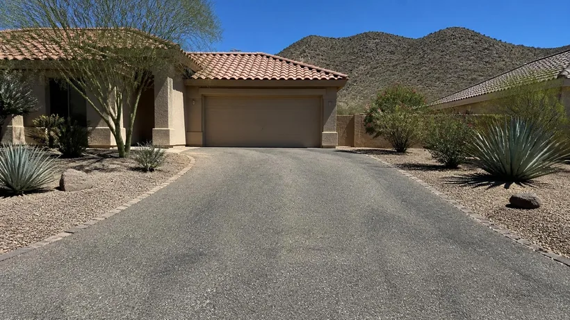 Asphalt driveway running to a tile-roofed stucco house past a desert gravel yard planted with agave