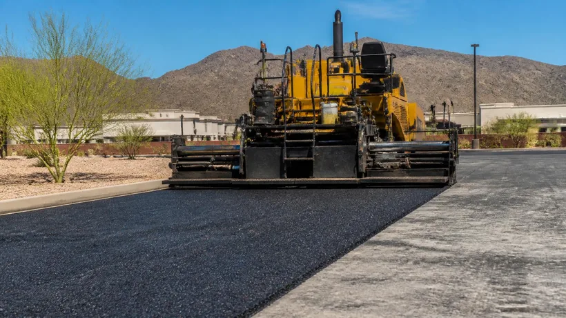Asphalt paver laying a fresh dark mat across a desert commercial lot with saguaro at the boundary